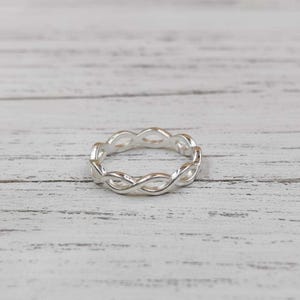 May include: A silver-colored ring with an infinity symbol design. The ring features a continuous pattern of interwoven loops, creating a delicate and elegant appearance. The ring is set against a white wooden surface.