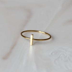 May include: A simple gold ring with a vertical bar accent. The ring is delicate and minimalist.