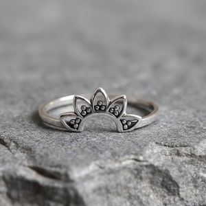 Op de afbeelding: Een zilverkleurige ring met een bloemmotief. De ring heeft een gebogen band met een reeks gestileerde bloemblaadjes. Elk bloemblad heeft een klein, gestippeld patroon. De ring ligt op een grijze, getextureerde ondergrond.