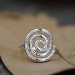 May include: A silver ring with a spiral design. The ring is made of a shiny, metallic material and is set against a neutral background. The spiral design is the main feature, creating a unique and eye-catching piece of jewellery.
