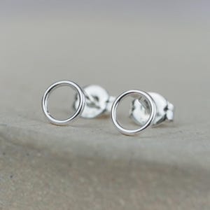 May include: A pair of small, silver-colored circle stud earrings. The earrings have a simple, minimalist design with a smooth, polished finish. The earrings are shown on a neutral background.
