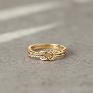 May include: A gold-colored ring with a knot design, adorned with small, sparkling gemstones. The ring features a double band and a knot-shaped centerpiece, creating a delicate and elegant look. The gemstones are set along the knot and the band, adding a touch of sparkle.