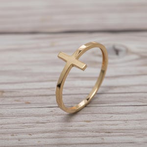 Könnte beinhalten: Ein goldfarbener Ring mit einem Kreuz-Design. Der Ring hat ein schlichtes Band mit einem Kreuz als Mittelpunkt. Der Ring wird auf einer hellen Holzoberfläche gezeigt.