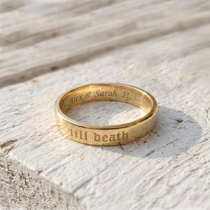 May include: A gold-toned metal band ring featuring the engraved text "till death" on the outer surface and the engraved text "Alex & Sarah 21. 11. 20" on the inner surface. The ring has a smooth, polished finish and a simple, circular design.