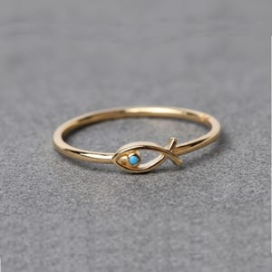 Op de afbeelding: Een goudkleurige ring met een vis-symbool en een kleine, ronde, turquoise steen. De ring heeft een delicate band en een gestileerd visontwerp, perfect voor dagelijks gebruik. Getoond op een grijze ondergrond.