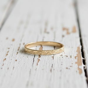Puede incluir: Un anillo dorado con un patrón de rayos de sol repetido. El anillo está sobre una superficie de madera blanca desgastada. El diseño del anillo presenta una serie de soles estilizados, añadiendo un toque de elegancia a la joya.