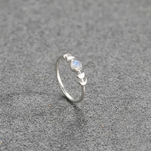 Puede incluir: Un anillo de plata con una piedra central redonda y clara, flanqueada por tres diseños de luna creciente. El anillo tiene una banda delgada y está sobre un fondo gris. El anillo es una joya.