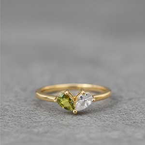 Puede incluir: Un anillo de oro con dos gemas en forma de pera, una verde y otra transparente. Las gemas están colocadas una al lado de la otra, creando un diseño único. El anillo está sobre una superficie gris.