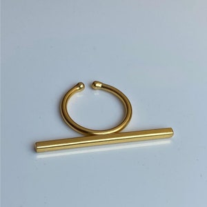 Puede incluir: Un anillo dorado con una barra recta que se desliza a través del anillo. El anillo está abierto y se puede ajustar para que se ajuste a diferentes tamaños de dedos.