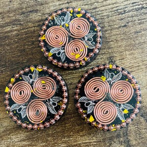 May include: Three round, black resin pieces with copper wire spirals, silver leaf accents, and a border of small copper beads. Yellow flecks are scattered around the edges. The pieces are arranged on a wooden surface.