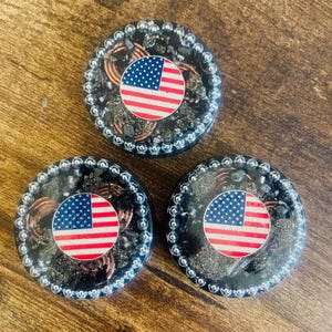 May include: Three round decorative objects with an American flag design. Each object features a circular flag with red and white stripes and a blue field of stars. The objects are surrounded by a dark, textured material and a silver-colored border.