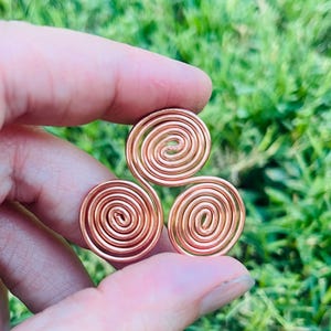 Op de afbeelding: Een koperkleurige hanger met drie spiralen. Elke spiraal is een cirkelvormige draadspoel, verbonden met de andere twee. De hanger wordt vastgehouden tegen een vage groene achtergrond van gras.