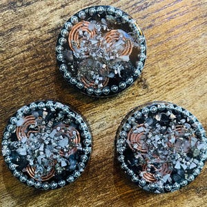 May include: Three round, resin-cast orgone energy generators. Each has a dark border with silver-colored beads, copper coils, and a mix of silver and black crystals. The objects are on a wooden surface.