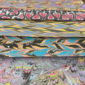 May include: Stack of five sheets of marbled paper with different patterns. The top sheet has a swirling pattern of purple, pink, and green. The second sheet has a pattern of black hearts on a white background. The third sheet has a pattern of yellow, blue, and black stripes. The fourth sheet has a pattern of blue, pink, and yellow swirls. The bottom sheet has a pattern of black, white, and yellow swirls.