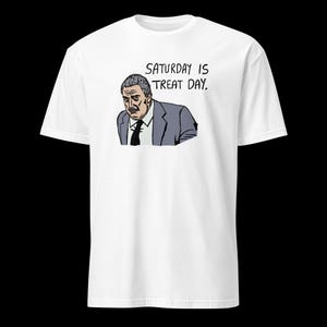 Puede incluir: Camiseta blanca con una ilustración de dibujos animados de un hombre con traje y el texto "SATURDAY IS TREAT DAY." La ilustración es en negro, gris y azul. La camiseta es de cuello redondo clásico.
