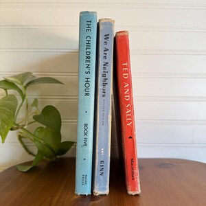 May include: Three vintage books with titles including "The Children's Hour," "We Are Neighbors," and "Ted and Sally." The books are in shades of blue and red, with visible text and wear. The books are standing on a wooden surface.