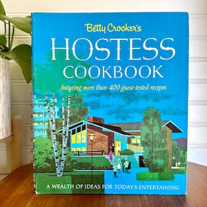 May include: A vintage cookbook titled "Betty Crocker's Hostess Cookbook" with a teal cover. The cover features a mid-century modern illustration of a house, trees, and people. The text reads "featuring more than 400 guest-tested recipes" and "A Wealth of Ideas for Today's Entertaining."