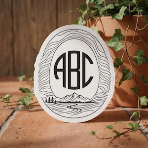 Può includere: Sottobicchiere ovale bianco con un monogramma nero delle lettere "ABC" all'interno di una cornice ovale. La cornice presenta un design ondulato e la parte inferiore raffigura una scena di montagna e fiume.