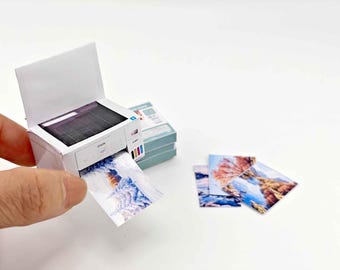 May include: A miniature white printer with a black top is printing a small picture. The printer has color ink cartridges on the side. Several printed pictures are next to the printer. The pictures have colorful landscape scenes.