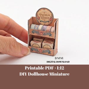 May include: A miniature cardboard display stand for a dollhouse, featuring two shelves stocked with tiny rolls of ribbon and small boxes. The top sign reads PREMIUM RIBBON. The unit is printed with floral motifs and wood grain patterns in brown tones.