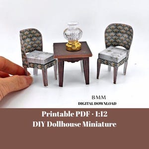 May include: Miniature dollhouse furniture set, including two chairs, a small table, and a decorative lamp. The chairs and table are brown with white legs. The chairs have patterned backs and seats. The lamp has a gold base and a clear glass shade. The text "Printable PDF - 1:12 DIY Dollhouse Miniature" is visible.