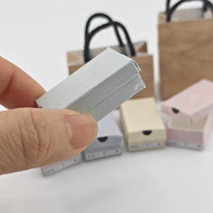 May include: A set of five miniature gift boxes, including a silver box with a black handle, a light pink box, a beige box, and two grey boxes. The boxes are all made of cardboard and have a simple design.