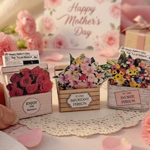 May include: Miniature wooden crates filled with colorful paper flowers, each with a unique message. One crate holds red roses, another a mix of blooms, and the third a variety of yellow and pink flowers. A "Happy Mother's Day" card is in the background.