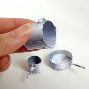 以下が含まれることがあります： 取っ手付きの銀色の紙製の鍋が3つあります。一番大きな鍋は手で持たれています。鍋はすべてサイズが異なります。