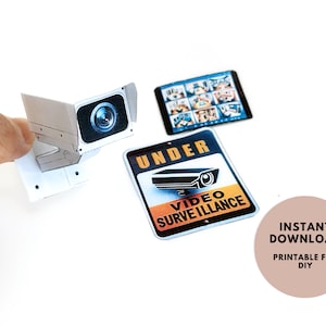 May include: Printable DIY security camera sign with the text "Under Video Surveillance". The sign is yellow, orange, and black with a white camera graphic. A small paper model of a security camera is also included.