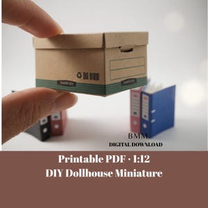 May include: A miniature brown cardboard Bankers Box with a green stripe and the text "BANKERS BOX". The box is held by a hand, with miniature binders in the background. The image also includes the text "Printable PDF 1:12 DIY Dollhouse Miniature".