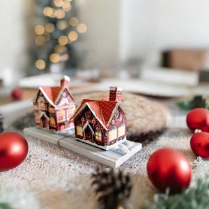 May include: Two miniature gingerbread houses with red roofs and white trim, decorated with candy and icing, sit on a white base. The houses are surrounded by red and white Christmas ornaments on a rustic, white, textured surface.