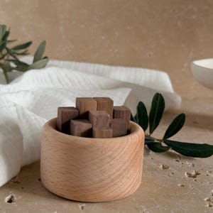 May include: A small, round, light-coloured wooden bowl containing a cluster of several dark brown wooden cubes. The bowl has a smooth, natural grain finish and a thick, rounded rim, while the cubes are arranged in a compact, stacked formation.