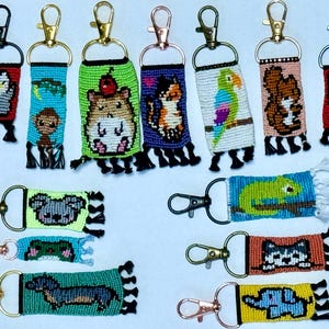 Può includere: Una collezione di portachiavi colorati con vari disegni di animali e natura. I portachiavi sono realizzati con piccole perline intrecciate insieme per creare immagini pixelate. Alcuni dei disegni includono un koala, un camaleonte, un pappagallo, un criceto, un gatto e un elefante.