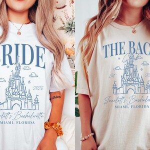 two women wearing white t-shirts with blue text and graphics. The text on the t-shirts reads "BRIDE" and "THE BACH" along with a castle illustration. The t-shirts also have the text "Seattle's Bachelorette" and "MIMI, FLORIDA" printed on them.