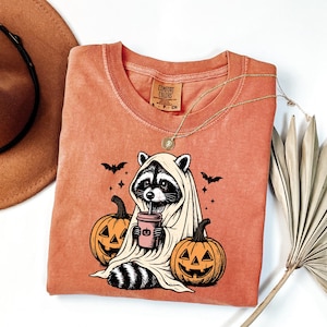 a Halloween-themed t-shirt with a raccoon wearing a ghost costume and holding a cup, surrounded by carved pumpkins and bats.