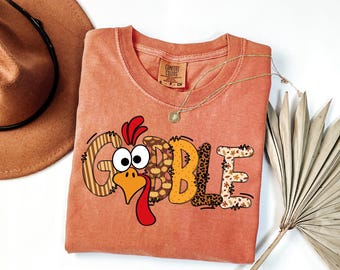 May include: A coral-colored t-shirt with the word "GOBBLE" in a turkey-themed design. The letters have various patterns, including stripes, leopard print, and fall leaves. A gold necklace and a brown hat are also visible.