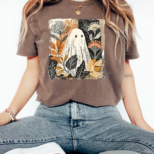 A woman wearing a brown t-shirt with a graphic of a ghost surrounded by flowers and leaves, sitting cross-legged on a white surface.