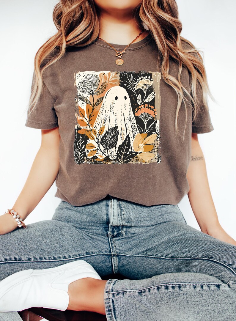 A woman wearing a brown t-shirt with a graphic of a ghost surrounded by flowers and leaves, sitting cross-legged on a white surface.