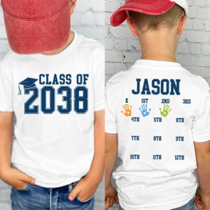 May include: White t-shirt with the text "CLASS OF 2038" in navy blue and a graduation cap graphic. The back of the shirt has the name "JASON" and handprint graphics indicating grade levels from kindergarten to 12th grade.