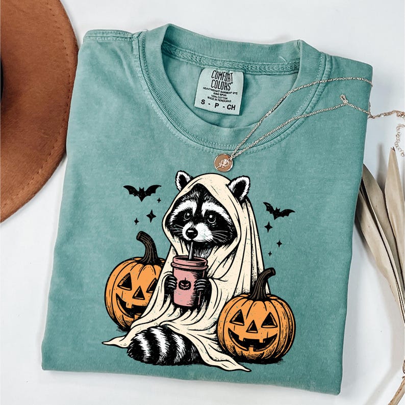 A t-shirt with a Halloween-themed graphic design, featuring a raccoon dressed as a ghost holding a coffee cup and two jack-o'-lanterns.