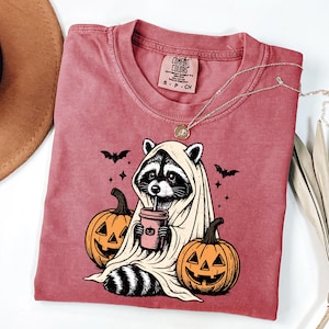A pink t-shirt with a graphic design depicting a raccoon dressed as a ghost, holding a coffee cup and surrounded by carved jack-o-lanterns.