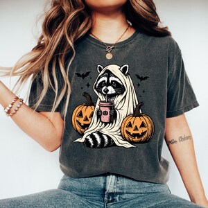 A woman wearing a t-shirt with a graphic design of a raccoon dressed as a ghost, holding a cup and surrounded by jack-o-lanterns.