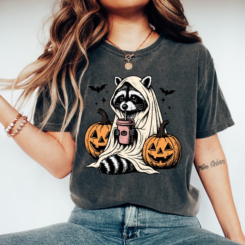 A woman wearing a t-shirt with a graphic design of a raccoon dressed as a ghost, holding a cup and surrounded by jack-o-lanterns.