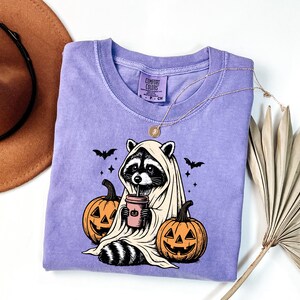 a purple t-shirt with a Halloween-themed graphic of a raccoon wearing a ghost costume and holding a pumpkin. The t-shirt is placed on a white surface, with a brown hat and a fan visible in the background.
