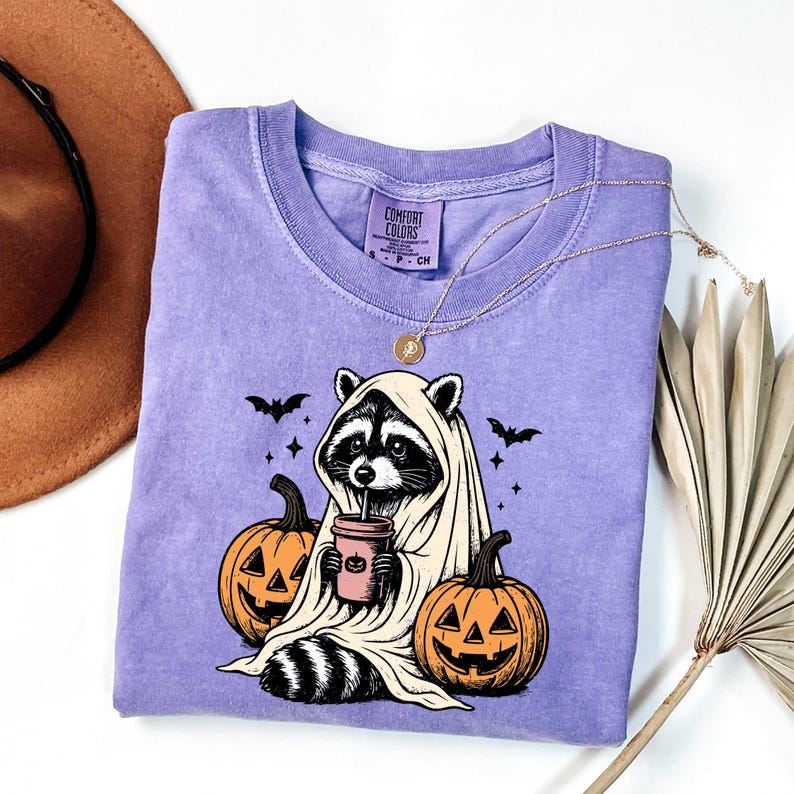 a purple t-shirt with a Halloween-themed graphic of a raccoon wearing a ghost costume and holding a pumpkin. The t-shirt is placed on a white surface, with a brown hat and a fan visible in the background.