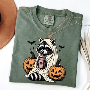 A green t-shirt with a Halloween-themed graphic design, including a raccoon wearing a ghost costume and holding a coffee cup, with jack-o-lanterns and bats around it.