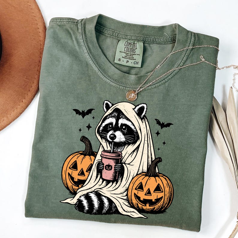 A green t-shirt with a Halloween-themed graphic design, including a raccoon wearing a ghost costume and holding a coffee cup, with jack-o-lanterns and bats around it.