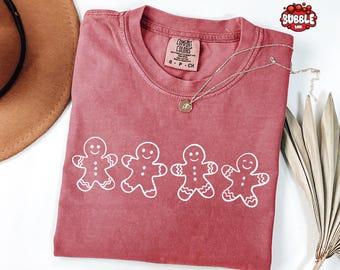 May include: A red-toned t-shirt with a crew neck and short sleeves. The shirt features a white graphic of four gingerbread men across the front. A silver necklace with a small pendant is draped on the shirt.