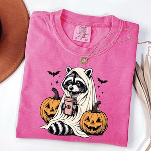 A pink t-shirt with a graphic design of a raccoon wearing a white sheet and holding a cup, surrounded by jack-o-lanterns, is displayed on a white surface alongside a brown hat and some dried leaves.