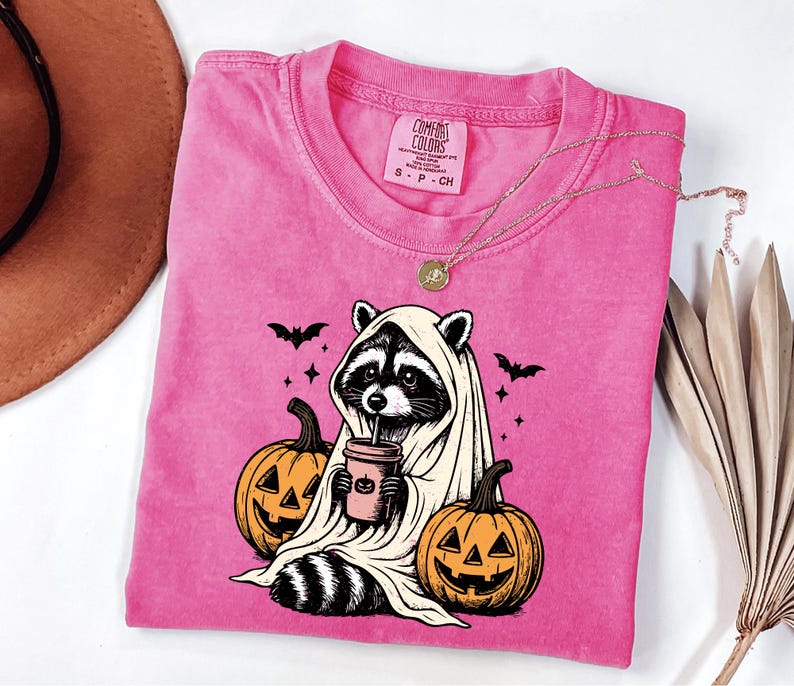 A pink t-shirt with a graphic design of a raccoon wearing a white sheet and holding a cup, surrounded by jack-o-lanterns, is displayed on a white surface alongside a brown hat and some dried leaves.